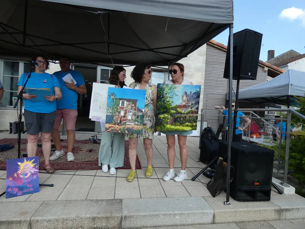 Remise des prix des concours devant la mairie de Rocheservière en présence de deux bénévoles et de trois femmes artistes qui montrent leurs tableaux au public