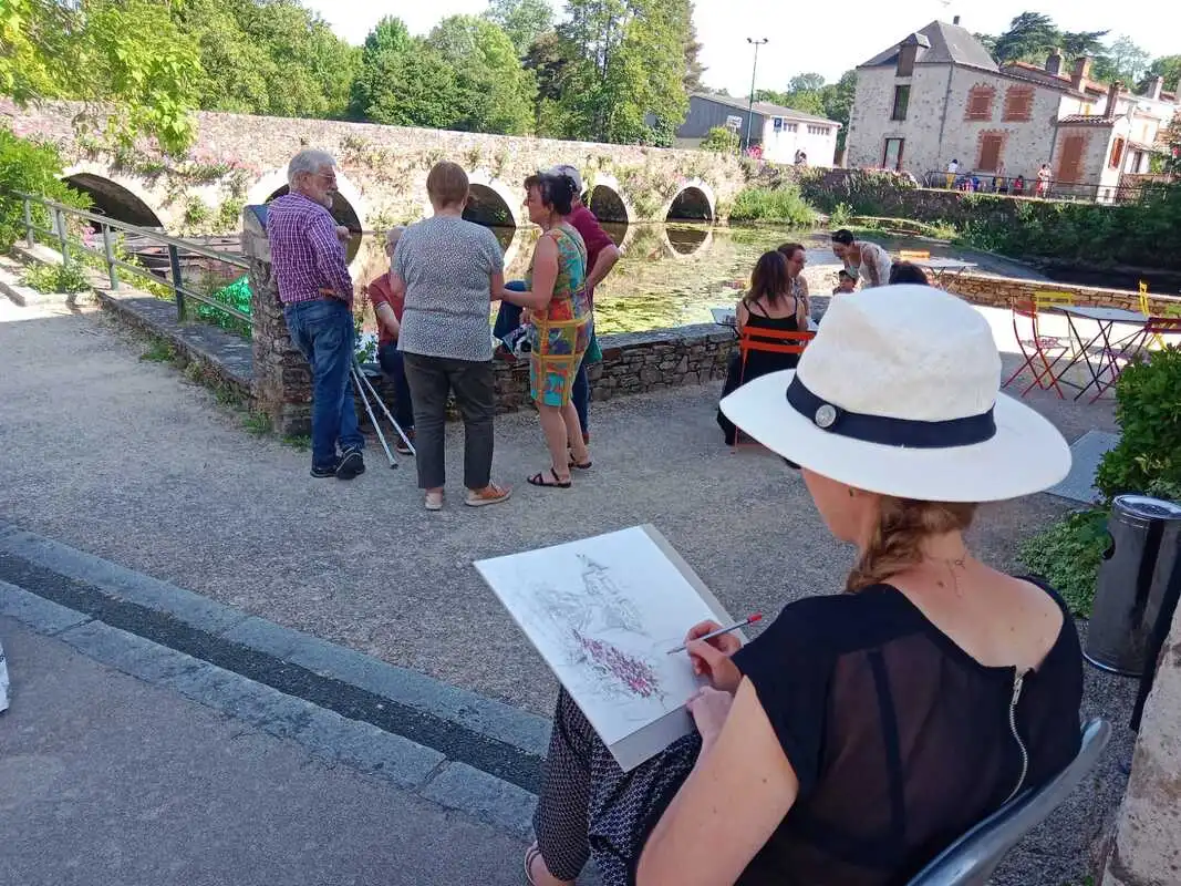 Femme qui dessine assise sur une chaise pendant le festival devant le vieux pont de la ville de Rocheservière et un groupe de personnes qui discutent